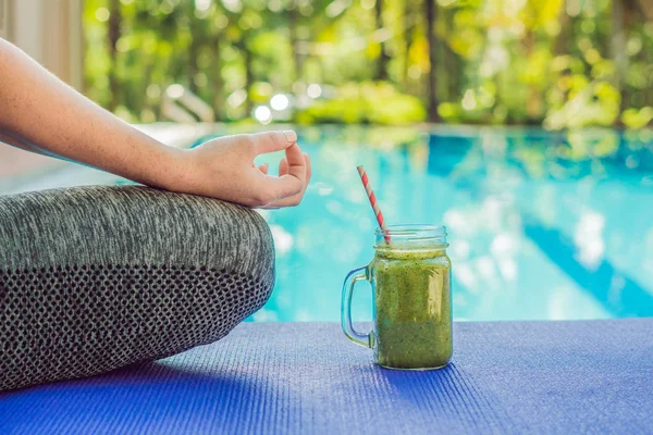 Closeup of a woman\'s hands during meditation with a green smoothies of spinach, orange and banana on the background of the pool.