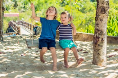 İki küçük sarışın erkek eğleniyor salıncakta tropikal kumlu sahilde.