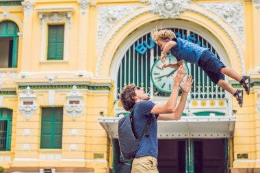 Father and son on background Saigon Central Post Office on blue sky background in Ho Chi Minh, Vietnam. Inscription in Vietnamese - Post Office. Steel structure of the gothic building was designed by Gustave Eiffel. Ho Chi Minh is a popular tourist d clipart