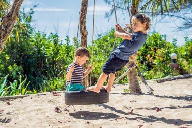 İki küçük sarışın erkek eğleniyor salıncakta tropikal kumlu sahilde.
