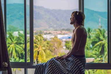 A man woke up in the morning and listens to music in wireless headphones on the background of a window overlooking the mountains. clipart