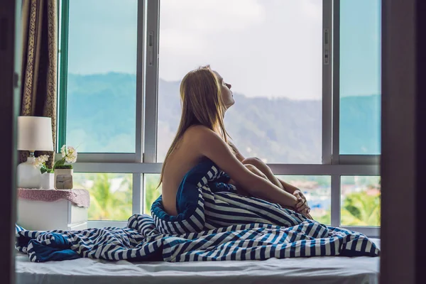 Young happy woman waking up in morning at bedroom, standing by window with beautiful mountain view