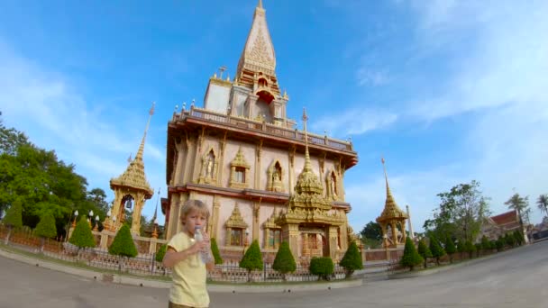 Foto en cámara lenta de un lindo niño bebiendo agua dulce de una botella frente a un templo budista de Wat Chalong en la isla de Phuket, Tailandia — Vídeos de Stock