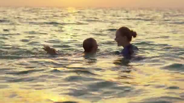 Une femme instructrice de natation enseigne à un petit garçon à nager dans une mer pendant un coucher de soleil — Video