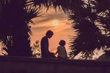 Baba ve oğlu, tropik fon ve palmiye ağaçlarının karşı günbatımı karşılamak siluetleri.