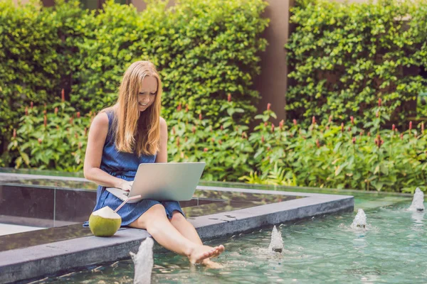 Young female freelancer sitting near pool with her laptop. Busy at holidays. Distant work concept