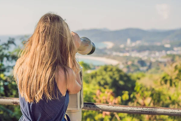 young beautiful blond woman enjoy the view with an coin operated binoculars. The water and the sky is blue. she wears a white dress and sunglasses. she feels good, is smiling and look to the horizon.