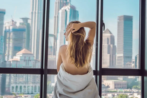 Young woman waking up in morning in apartment in downtown area with view of the skyscrapers. Life in noise of big city concept. Not enough sleep.