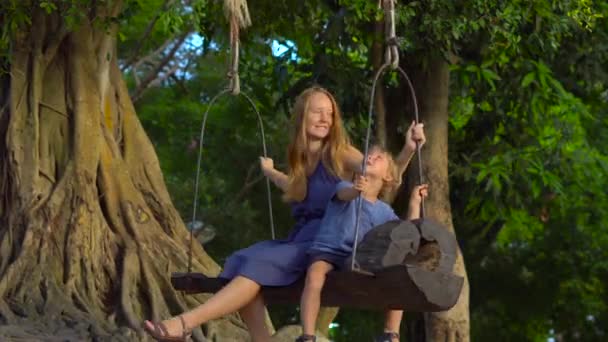 母と息子は、熱帯公園に大きな熱帯の木に掛かっているエキゾチックな古いスイングでスイングします。子供の概念と旅行します。 — ストック動画