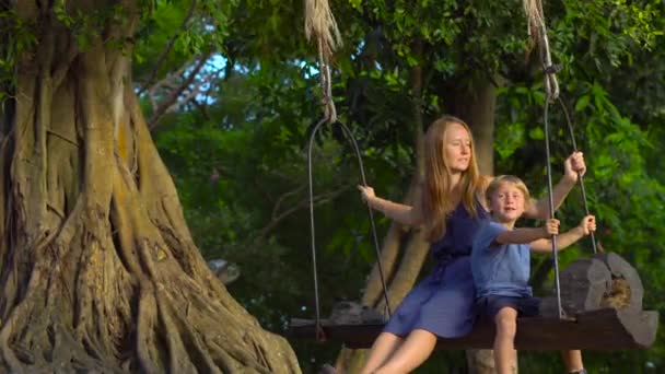 Mãe e filho balançam em um velho baloiço exótico pendurado em uma grande árvore tropical em um parque tropical. Viagem com conceito de crianças — Vídeo de Stock