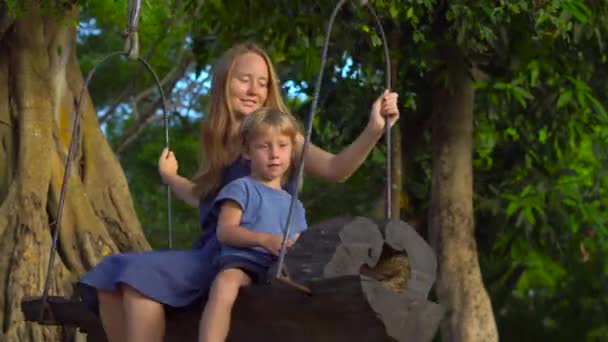 Madre e figlio dondolano su una vecchia altalena esotica appesa ad un grande albero tropicale in un parco tropicale. Viaggiare con i bambini concetto — Video Stock