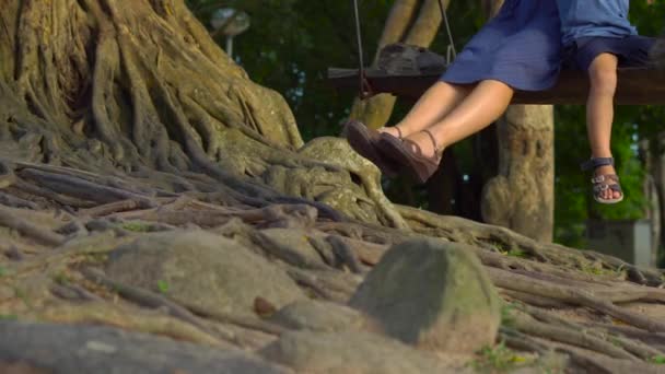 Madre e figlio dondolano su una vecchia altalena esotica appesa ad un grande albero tropicale in un parco tropicale. Viaggiare con i bambini concetto — Video Stock