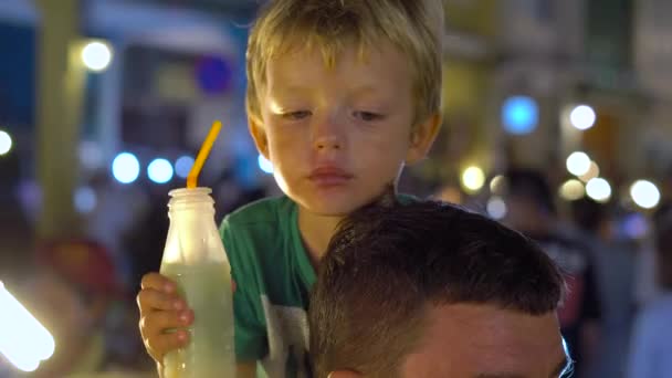 Маленький хлопчик вживаю свіжі фруктові соки сидить на шиї батьків на нічний ринок в Таїланді. Концепція тайської кухні. Азіатський харчової концепції — стокове відео