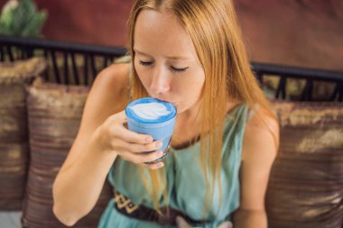 Akdeniz kahvaltısı yapan genç bir kadın kanepede oturuyor ve Moda içeceği olan mavi latte içiyor. Sıcak kelebek bezelye latte veya mavi spirulina latte.