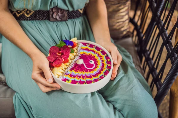 Mujer joven tomando un desayuno mediterráneo sentada en su sofá y come desayuno tropical saludable, batido con frutas tropicales, decorado con un patrón de yogur colorido con cúrcuma y — Foto de Stock