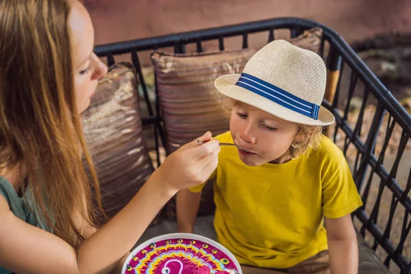 Mother and son having a mediterranean breakfast seated at the sofa and eats Healthy tropical breakfast, smoothie bowl with tropical fruits, decorated with a pattern of colorful yogurt with turmeric — Stock Photo, Image