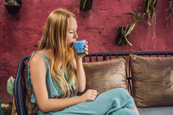 Молодая женщина, завтракающая в средиземноморском стиле, сидит у дивана и пьет модный напиток: синий латте. Горячий латте с горохом или голубой спирулиновый латте — стоковое фото