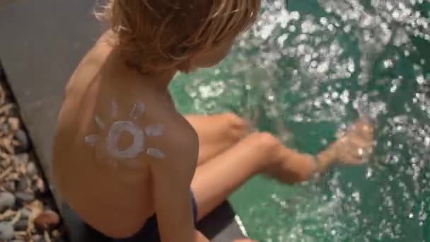 Zeitlupenaufnahme eines kleinen Jungen mit einer Sonne, die von einer Sonnencreme auf seiner Schulter gezeichnet wird, spritzt Wasser in einem Schwimmbad. Sonnenschutzkonzept — Stockvideo