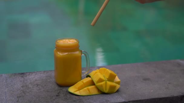 Primer plano de un vaso de un batido de mango y mangos cortados en un lado de una piscina con una paja de bambú bebiendo en ella. Alternativa a las pajitas de plástico. Concepto de productos ecológicos — Vídeos de Stock
