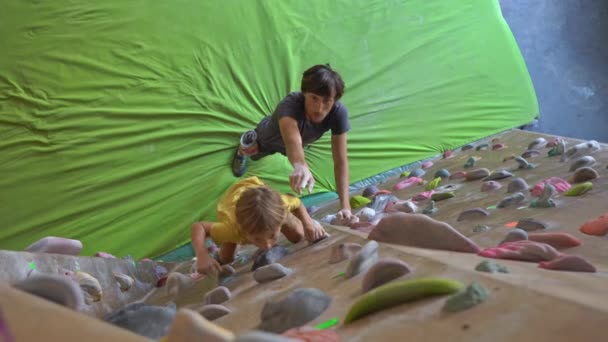 Un joven instructor de escalada enseñando a un niño a escalar la pared en un gimnasio de escalada — Vídeos de Stock