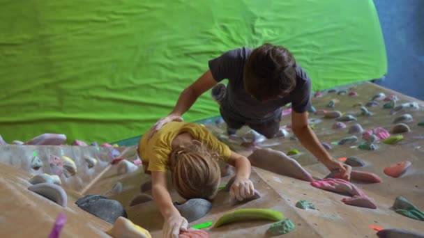 A young man climbing instructor teaching little boy how to climb the wall in a bouldering climbing gym — Stock Video