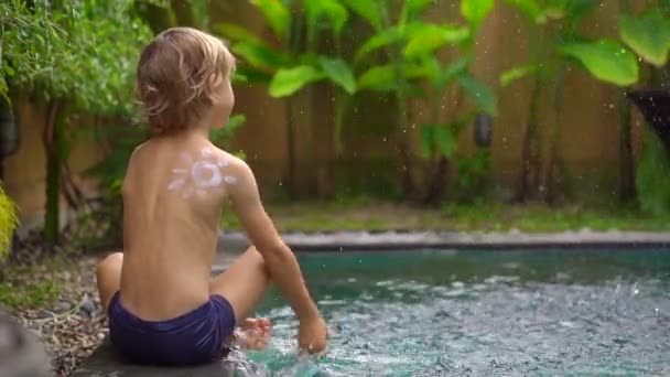 Lassított felvétel egy kisfiúról, akit naptejszínnel rajzolt a vállára, és vizet fröcskölt egy medencébe. Napvédelmi koncepció — Stock videók