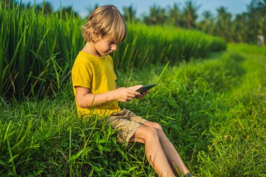 Sahada tablet tutan mutlu çocuk. Güneşli bir günde çimenlerin üzerinde oturan çocuk. Evde eğitim veya tablet oynama
