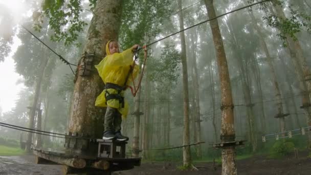 Caméra d'action prise d'un petit garçon dans un harnais de sécurité grimpe sur un chemin dans la cime des arbres dans un parc d'aventure forestière. Il grimpe sur le sentier des hautes cordes. Centre de loisirs extérieur avec activités d'escalade — Video