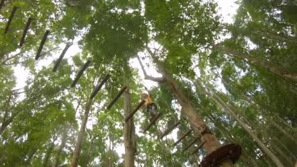 La cámara de acción de un niño pequeño con un arnés de seguridad sube a una ruta en las copas de los árboles en un parque de aventuras forestales. Se sube a un sendero de cuerda alta. Centro de diversiones al aire libre con actividades de escalada — Vídeo de stock