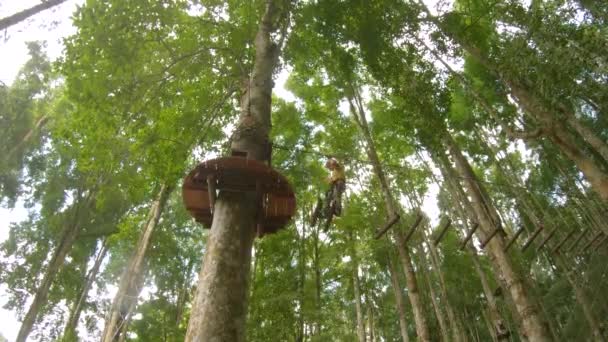 La cámara de acción de un niño pequeño con un arnés de seguridad sube a una ruta en las copas de los árboles en un parque de aventuras forestales. Se sube a un sendero de cuerda alta. Centro de diversiones al aire libre con actividades de escalada — Vídeos de Stock