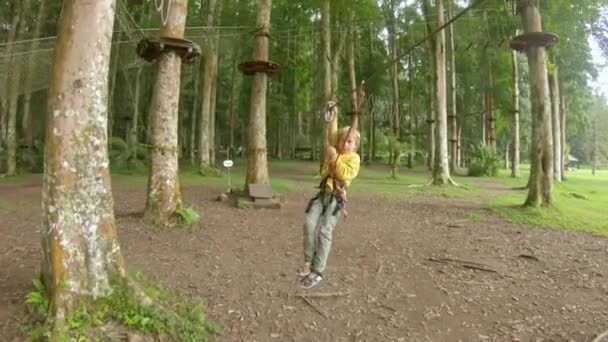 La cámara de acción de un niño pequeño con un arnés de seguridad sube a una ruta en las copas de los árboles en un parque de aventuras forestales. Se sube a un sendero de cuerda alta. Centro de diversiones al aire libre con actividades de escalada — Vídeos de Stock