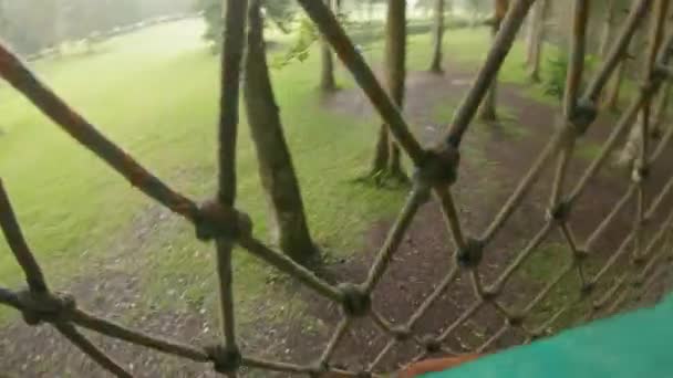 Punto de vista plano de un hombre en un arnés de seguridad que sube a una ruta en las copas de los árboles en un parque de aventura forestal. Se sube a un sendero de cuerda alta. Centro de diversiones al aire libre con actividades de escalada — Vídeos de Stock