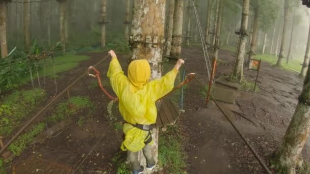 Action camera shot of a little boy in a safety harness climbs on a route in treetops in a forest adventure park. He climbs on high rope trail. Outdoor amusement center with climbing activities — Stock Video