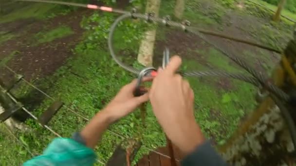 Punto de vista plano de un hombre en un arnés de seguridad que sube a una ruta en las copas de los árboles en un parque de aventura forestal. Se sube a un sendero de cuerda alta. Centro de diversiones al aire libre con actividades de escalada — Vídeos de Stock