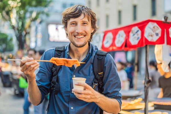 Young man tourist eating Typical Korean street food on a walking street of Seoul. Spicy fast food simply found at local Korean martket, Soul Korea — Stock Photo, Image