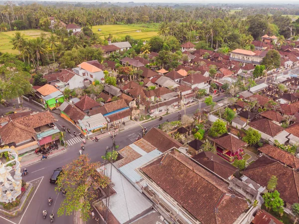 Many villas with brown-orange shingle roofs between tropical trees on the sky background in Ubud on Bali. Sun is shining onto them. Aerial horizontal photo