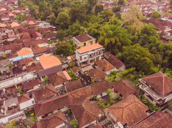 Many villas with brown-orange shingle roofs between tropical trees on the sky background in Ubud on Bali. Sun is shining onto them. Aerial horizontal photo