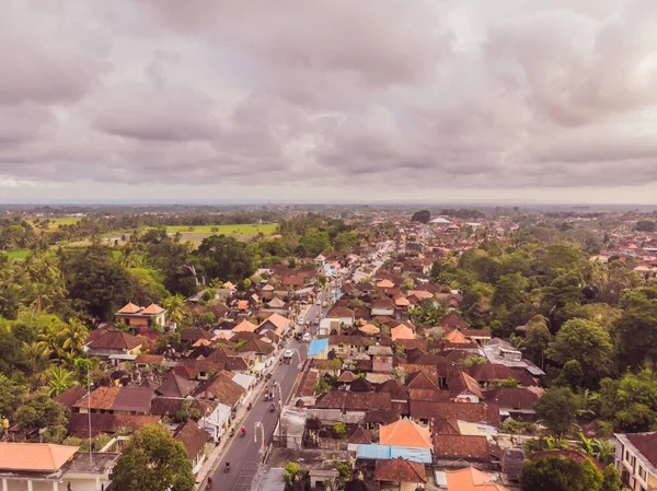 Many villas with brown-orange shingle roofs between tropical trees on the sky background in Ubud on Bali. Sun is shining onto them. Aerial horizontal photo