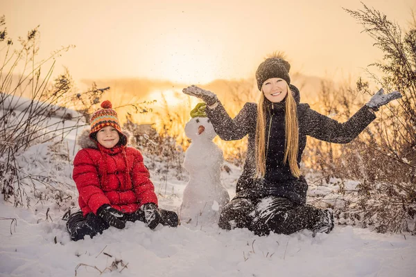Счастливая семья в теплой одежде. Улыбающиеся мать и сын делают снеговика на открытом воздухе. Концепция зимних мероприятий — стоковое фото