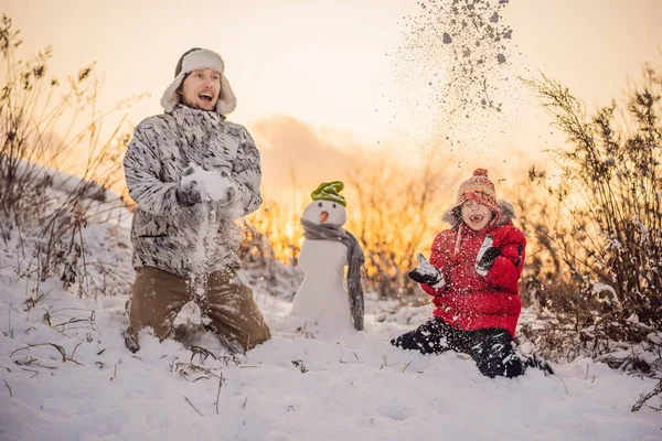 Boldog család meleg ruhában. Mosolygó apa és fia hóembert csinálnak a szabadban. A téli tevékenységek fogalma — Stock Fotó