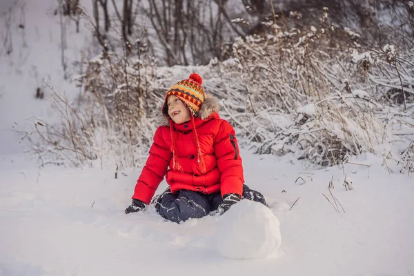 Rapaz bonito em roupas vermelhas de inverno constrói um boneco de neve. Inverno divertido conceito ao ar livre — Fotografia de Stock