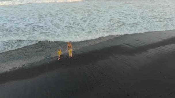 Luchtfoto van een moeder en haar zoon wandelen en plezier hebben op een strand met een zwarte vulkanische zand — Stockvideo