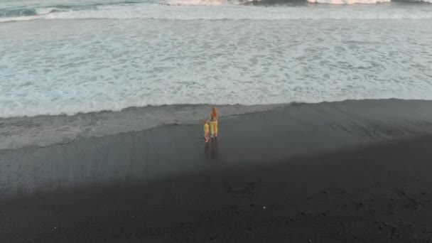 Foto aérea de una madre y su hijo caminando y divirtiéndose en una playa con arena volcánica negra — Vídeos de Stock