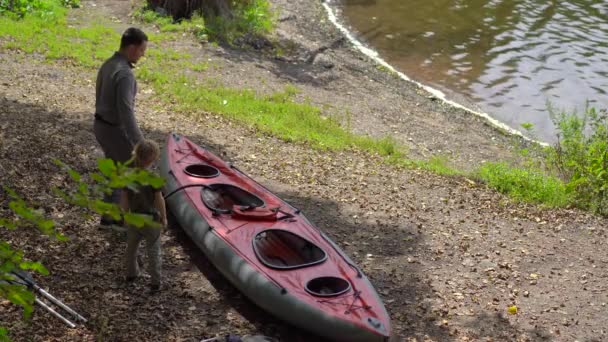 Молодой человек и его маленький сын на озере или на берегу моря готовят большой надувной каяк для купания — стоковое видео