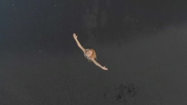 Siyah bir volkanik kumla sahilde yürüyen ve eğlenen genç bir kadının havadan çekilmiş fotoğrafı.