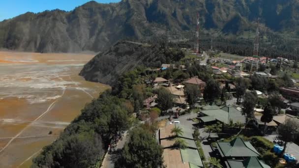 Luftaufnahme des berühmten aktiven Bromo-Vulkans oder des Gunung Bromo und Batok-Vulkans innerhalb der Tengger Caldera auf der Java-Insel. Drohne fliegt am Rande der Caldera, wo das Dorf liegt — Stockvideo