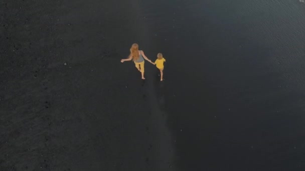Bir anne ve oğlunun siyah bir volkanik kumla kumsalda yürüyüp eğlenirken çekilmiş bir fotoğrafı. — Stok video