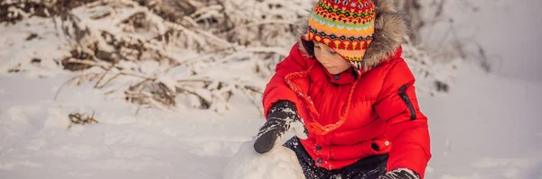 Söt pojke i röda vinterkläder bygger en snögubbe. Vinternöje utomhus koncept Banner, Long Format — Stockfoto