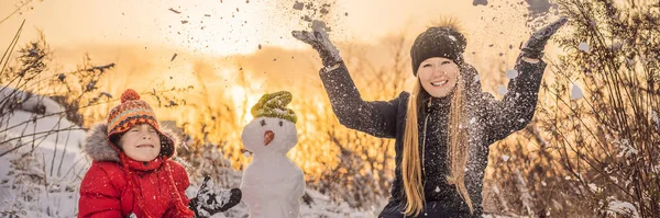 穿着保暖衣服的快乐家庭。 笑着妈妈和儿子在外面堆雪人 冬季活动的概念，横幅，长格式 — 图库照片