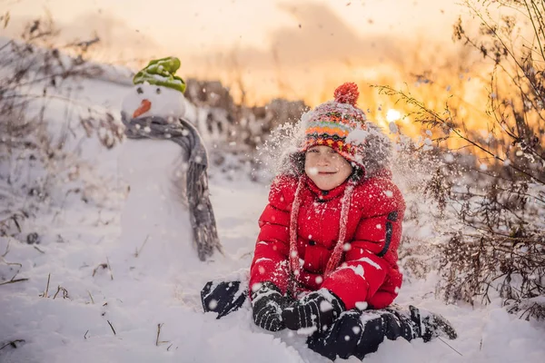 Aranyos fiú piros téli ruhákban épít egy hóembert. Téli szórakozás Kültéri koncepció — Stock Fotó